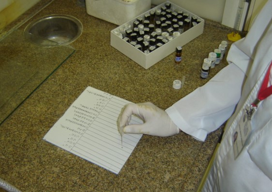 Em uma bancada de mármore aparece uma mão com luva colocando gotas de alguma substância em um papel. Ao lado, há uma caixa cheia de vidros escuros com substâncias.